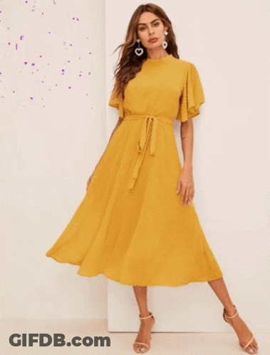 A Woman Wearing A Yellow Dress And Earrings Stands In Front Of A White Wall GIF