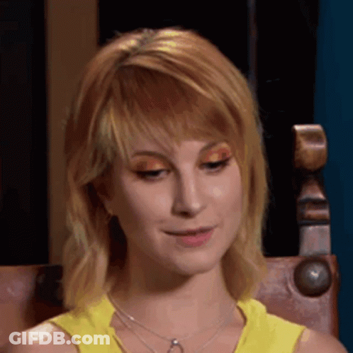 A Woman Wearing A Yellow Tank Top And A Necklace Is Sitting In A Chair And Smiling . GIF