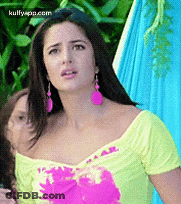 A Woman Wearing A Yellow Top And Pink Earrings Is Standing In Front Of A Blue Hammock . GIF