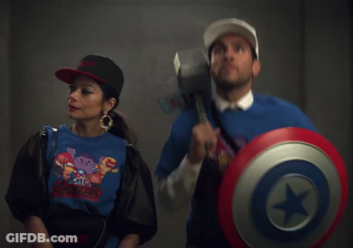 A Woman Wearing An Avengers Shirt Stands Next To A Man Holding A Shield And Hammer GIF