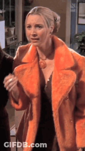 A Woman Wearing An Orange Fur Coat Is Standing In A Room With Other People . GIF