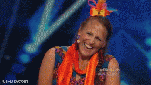 A Woman Wearing An Orange Scarf And A Colorful Hat Is Laughing On A Stage . GIF