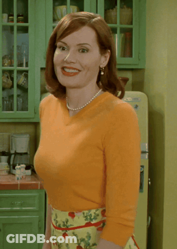 A Woman Wearing An Orange Top And A Yellow Skirt Smiles In A Kitchen GIF