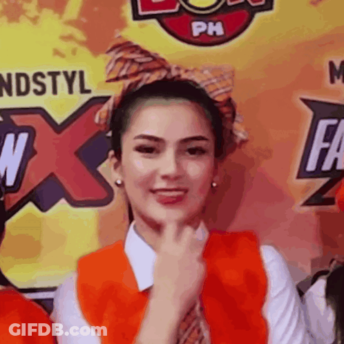 A Woman Wearing An Orange Vest And Tie Stands In Front Of A Ph Logo GIF