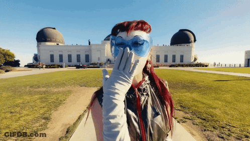 A Woman Wearing Blue Glasses And White Gloves Covering Her Face In Front Of A Building GIF