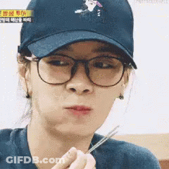 A Woman Wearing Glasses And A Baseball Cap Is Holding Chopsticks In Her Hand . GIF