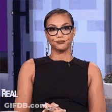 A Woman Wearing Glasses And A Black Dress Is Sitting On A Couch . GIF