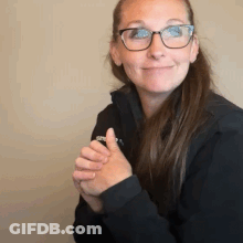A Woman Wearing Glasses And A Black Jacket Is Smiling While Holding Her Hands Together . GIF