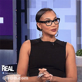 A Woman Wearing Glasses And A Black Tank Top Is Sitting In Front Of A Sign That Says Real GIF