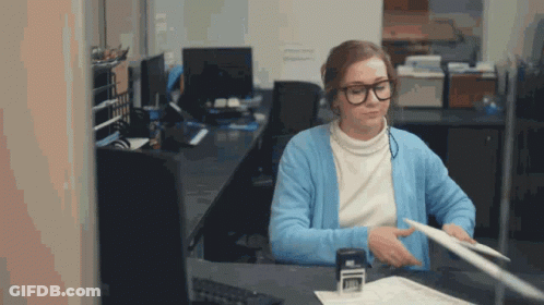 A Woman Wearing Glasses And A Blue Cardigan Is Sitting At A Desk GIF