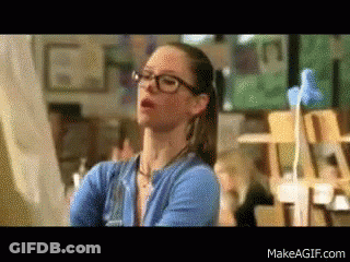A Woman Wearing Glasses And A Blue Sweater Is Standing In Front Of A Painting GIF