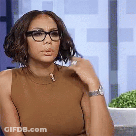 A Woman Wearing Glasses And A Brown Tank Top Is Sitting Down And Looking At The Camera . GIF