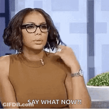 A Woman Wearing Glasses And A Brown Tank Top Is Sitting Down And Saying `` Say What Now '' . GIF
