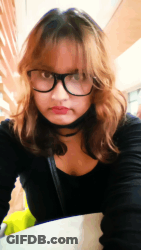 A Woman Wearing Glasses And A Choker Is Sitting At A Table GIF