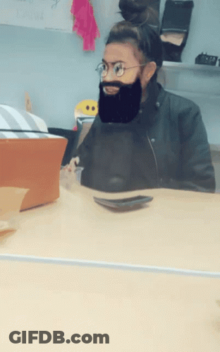 A Woman Wearing Glasses And A Fake Beard Sits At A Table GIF