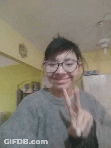 A Woman Wearing Glasses And A Gray Jacket Is Making A Peace Sign GIF