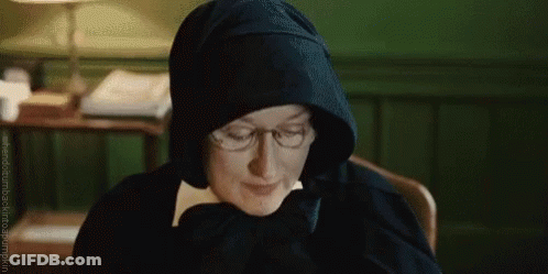 A Woman Wearing Glasses And A Hood Is Sitting At A Desk . GIF