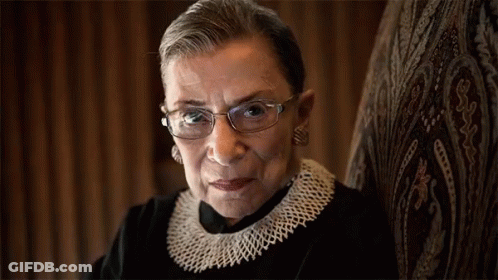 A Woman Wearing Glasses And A Knitted Collar Looks At The Camera GIF