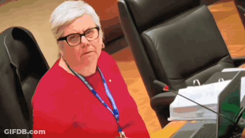 A Woman Wearing Glasses And A Lanyard That Says ' A ' On It Sits In Front Of A Laptop GIF