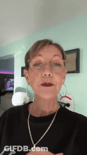 A Woman Wearing Glasses And A Necklace Looks At The Camera While Making A Face GIF