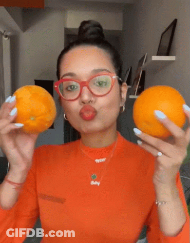 A Woman Wearing Glasses And A Necklace That Says Baby Holds Two Oranges GIF
