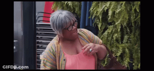 A Woman Wearing Glasses And A Pink Shirt Is Standing In Front Of A Stack Of Chairs . GIF