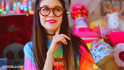 A Woman Wearing Glasses And A Tie Dye Shirt Is Standing In Front Of A Pile Of Stuffed Animals . GIF