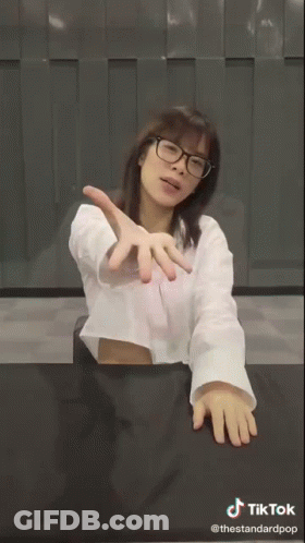 A Woman Wearing Glasses And A White Shirt Is Sitting At A Table With A Sign That Says Wee GIF
