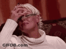 A Woman Wearing Glasses And A White Sweater Is Sitting On A Couch And Covering Her Face With Her Hand . GIF
