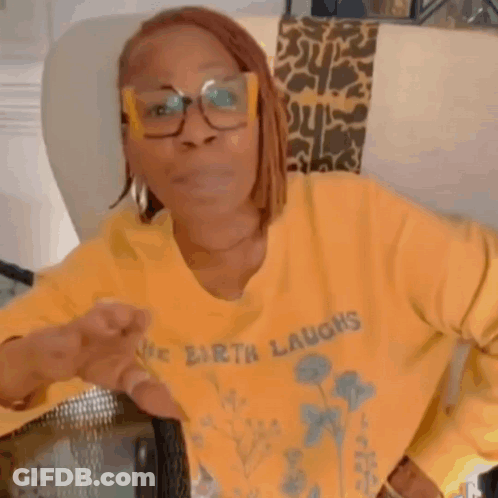 A Woman Wearing Glasses And A Yellow Sweater Is Sitting In A Chair And Making A Funny Face . GIF