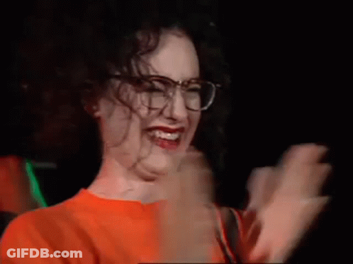 A Woman Wearing Glasses And An Orange Shirt Is Laughing In A Dark Room . GIF