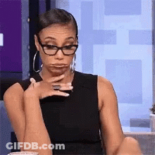 A Woman Wearing Glasses And Hoop Earrings Is Sitting At A Table Making A Funny Face . GIF