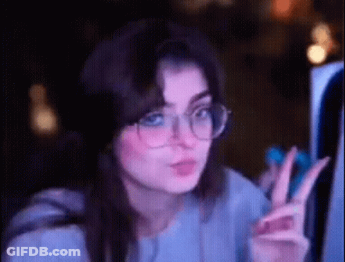 A Woman Wearing Glasses Is Giving A Peace Sign While Looking At A Computer Screen . GIF