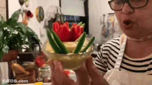 A Woman Wearing Glasses Is Holding A Glass Filled With Vegetables And Flowers . GIF