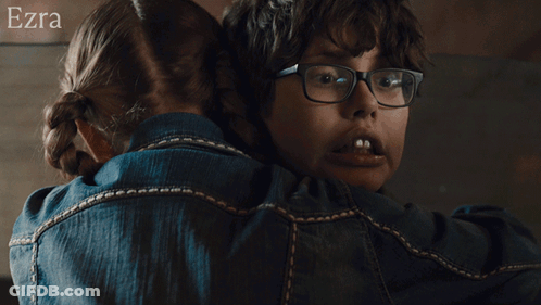 A Woman Wearing Glasses Is Hugging Another Woman Wearing A Blue Shirt With The Name Ezra On The Bottom GIF