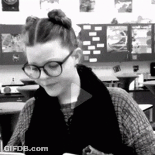 A Woman Wearing Glasses Is Sitting At A Desk In A Classroom . GIF