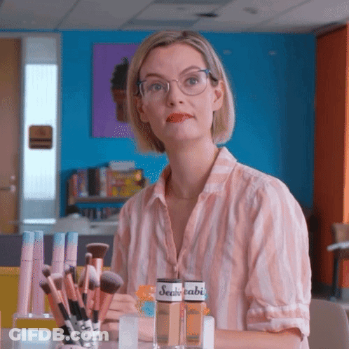 A Woman Wearing Glasses Is Sitting At A Table With Makeup Brushes And A Bottle Of Sealedabi GIF