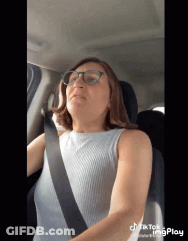 A Woman Wearing Glasses Is Sitting In A Car With A Seat Belt Around Her Neck GIF