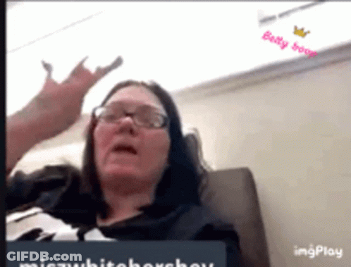 A Woman Wearing Glasses Is Sitting On A Couch And Making A Devil Horns Gesture . GIF