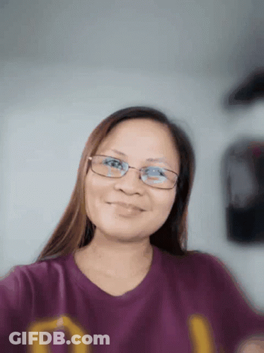 A Woman Wearing Glasses Is Smiling And Wearing A Purple Shirt With The Letter D On It GIF