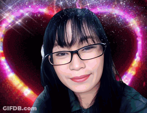 A Woman Wearing Glasses Is Smiling In Front Of A Heart GIF