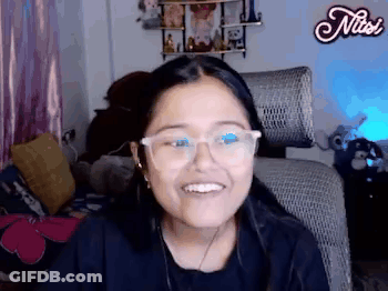 A Woman Wearing Glasses Is Smiling While Sitting In A Chair In A Room . GIF