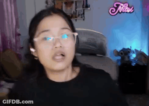 A Woman Wearing Glasses Is Standing In Front Of A Pink Neon Sign That Says Nioi GIF