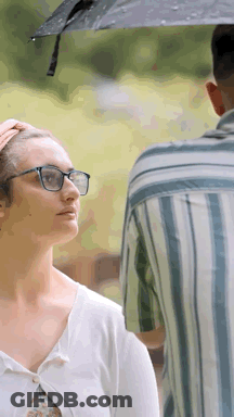 A Woman Wearing Glasses Is Standing Next To A Man Holding An Umbrella Over Her Head . GIF