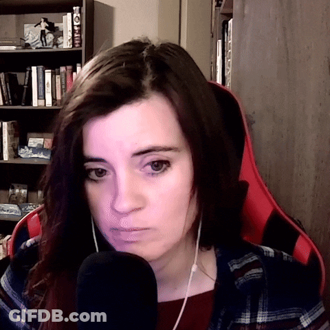 A Woman Wearing Headphones And A Plaid Shirt Sits In Front Of A Microphone With A Bookshelf In The Background GIF