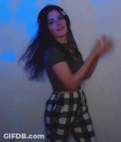 A Woman Wearing Headphones And Plaid Pants Is Dancing In Front Of A Blue Wall . GIF