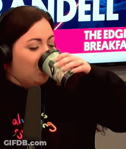 A Woman Wearing Headphones Drinks From A Cup In Front Of A Screen That Says " The Edge Breakfast " GIF