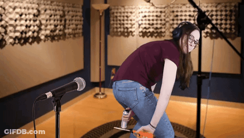 A Woman Wearing Headphones Is Kneeling In Front Of A Microphone And Holding A Can Of Elmer 'S Glue GIF