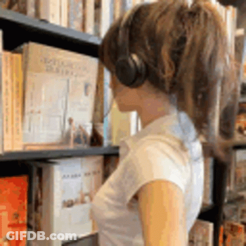 A Woman Wearing Headphones Is Looking At Books In A Library GIF