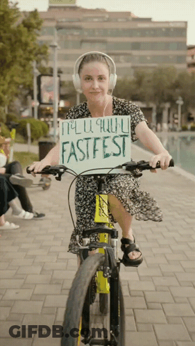 A Woman Wearing Headphones Is Riding A Bike While Holding A Sign That Says Fastfest GIF